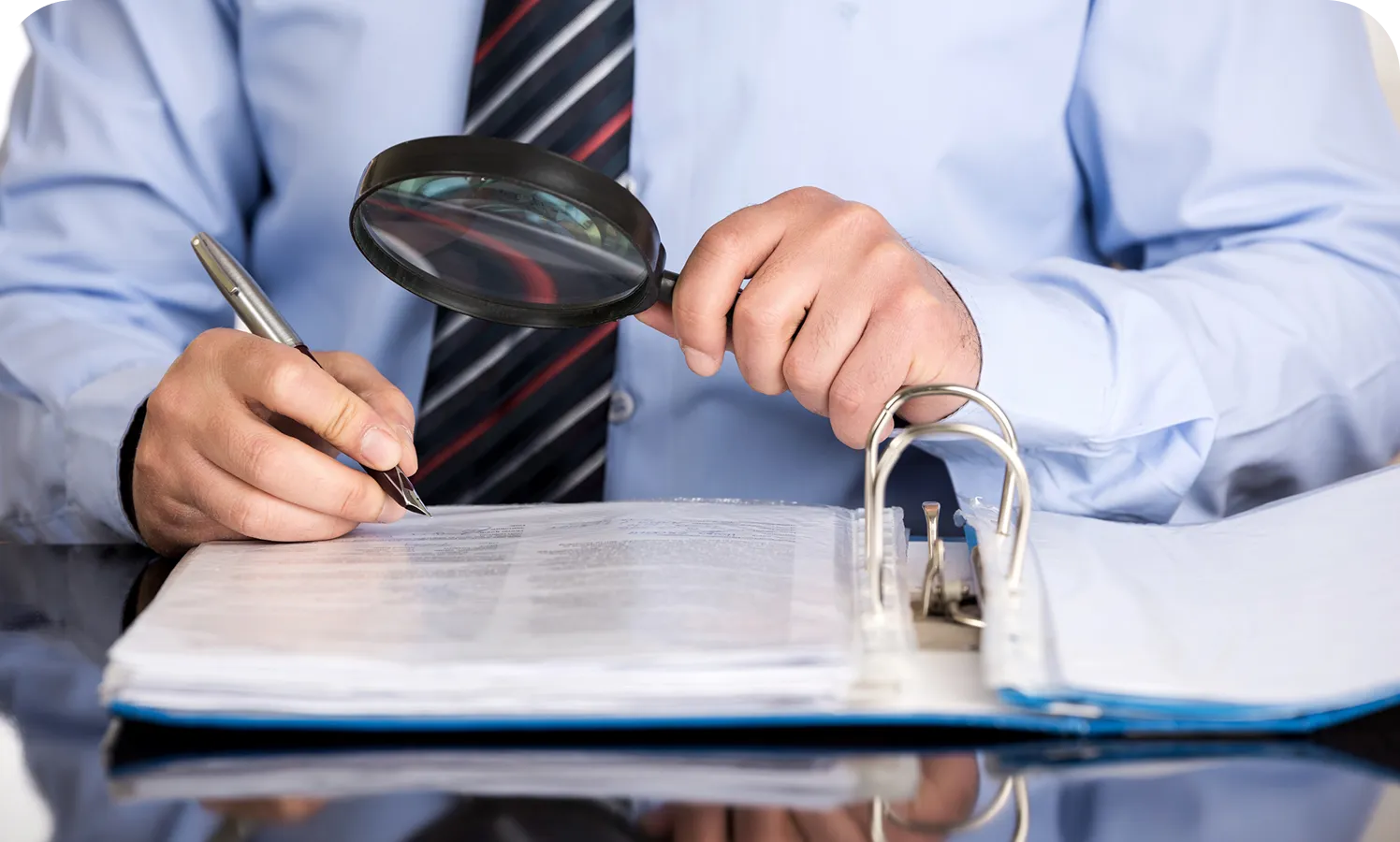 Grayson Risk Consulting Person in a blue shirt and tie holding a magnifying glass, reading a document in a binder, and writing with a pen.